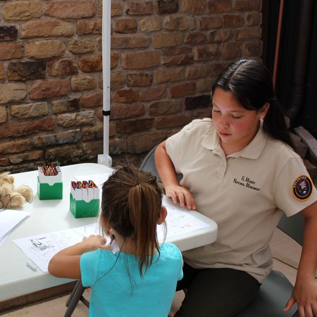 A young volunteer oversees a young girl at a table full of cayons and markers.
