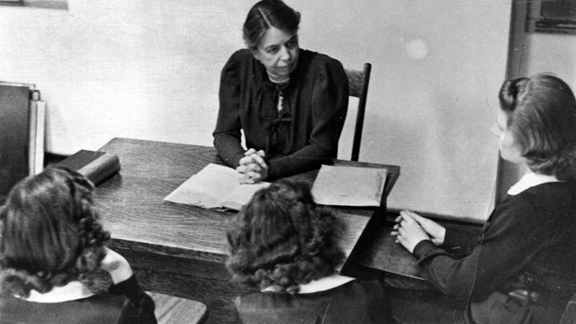 A woman at a desk conducting a class with three girls.