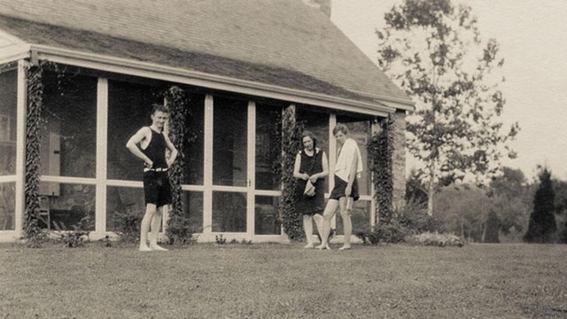 A man and two women in bathing suits stand before the stone cottage.