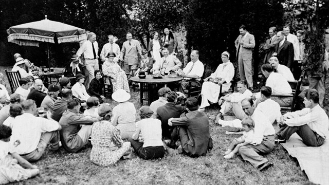 A large group of people outdoors, gathered around a table while a man reads to them.
