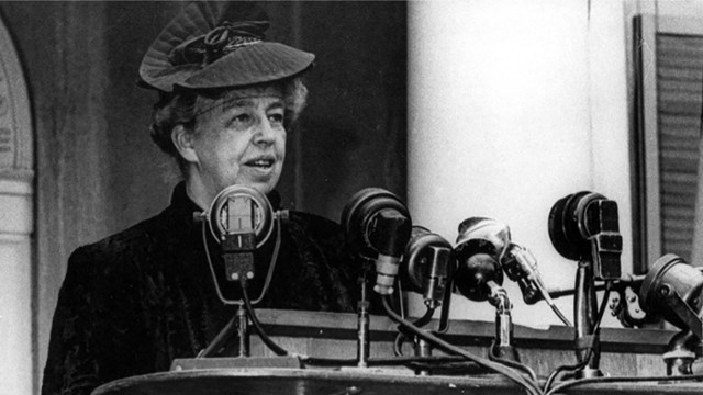 A woman wearing a fur coat and hat delivering a speech behind a podium with microphones.