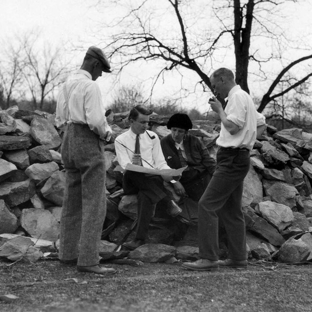 Three men and a women seated by a pile of stone examine a drawing.