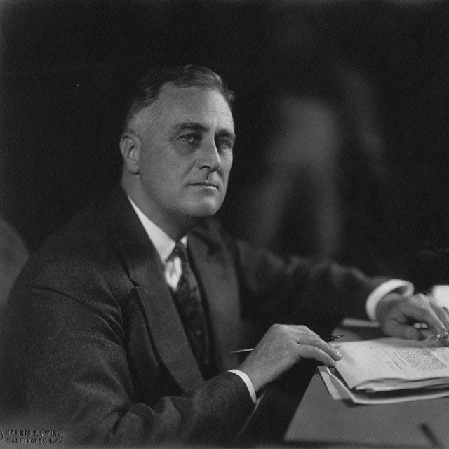 A man in a dark suit seated behind a desk with papers.