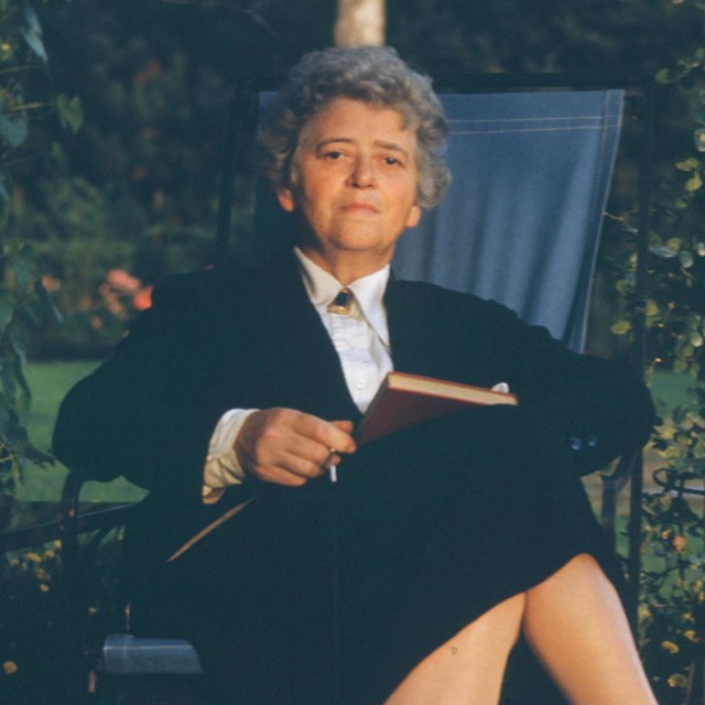 A woman in a dark dress seated outdoors with a book.