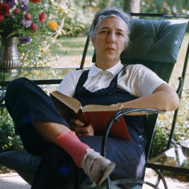 A woman in overalls seated outdoors with a book.