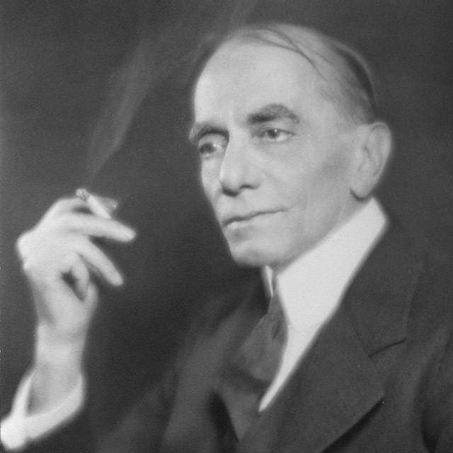 A man in a dark suit holding a cigarette.