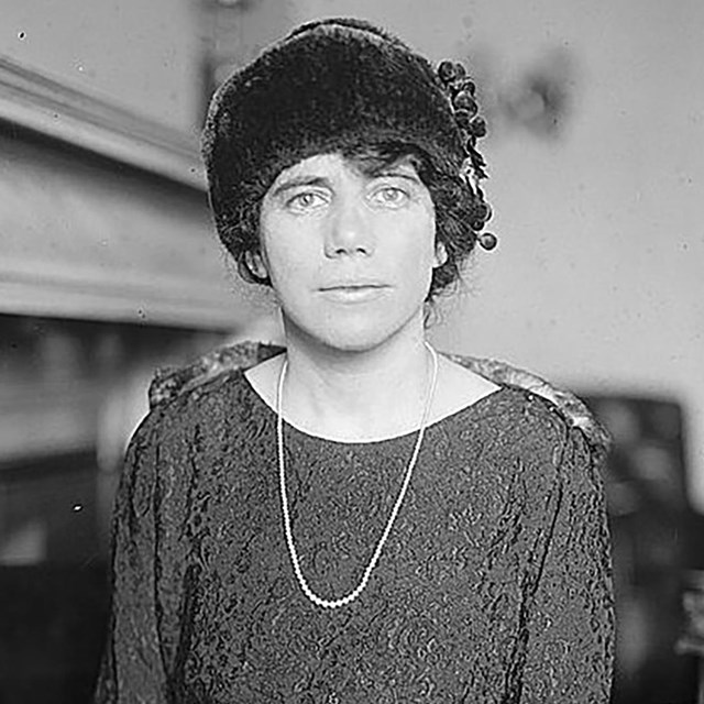 A seated woman wearing a dress and dark hat.