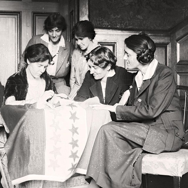 A group of women gathered around a table sewing a flag.