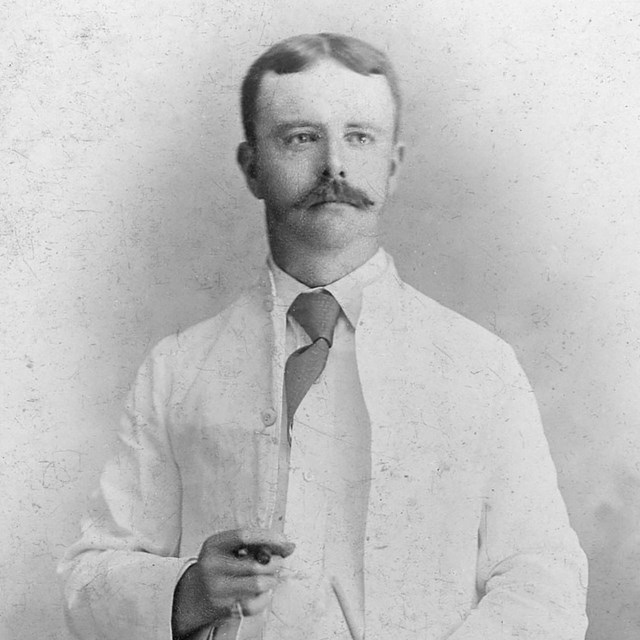 A man wearing a white suit, holding a hat and a cigar.
