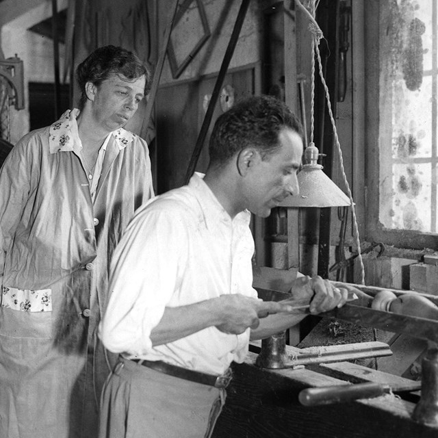 A woman in a factory looks over the shoulder of a man turning wood on a lathe.