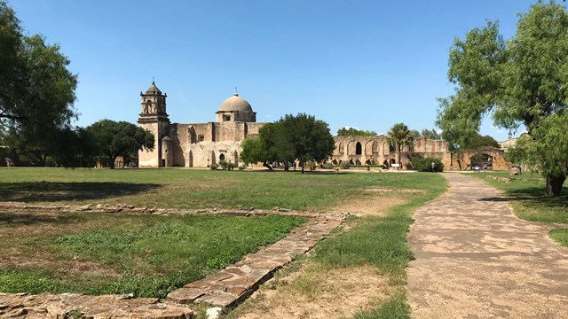 A spanish-colonial style mission in the background of a lawn with trees.