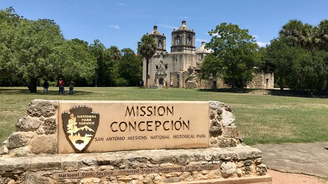 A stone sign stands in front of a tall, weathered, spanish-colonial style mission.