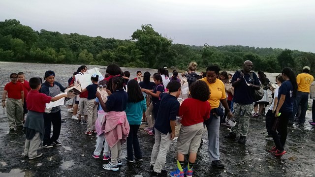 A group of young students and adults stand near a river on a cloudy day.