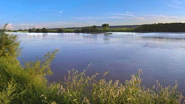 Un río ancho y tranquilo corre entre riberas con vegetación verde y baja.