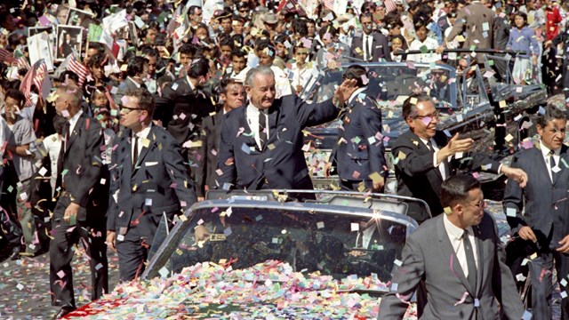 Presidentes se paran y saludan de coche en desfile. Multitud alinea la calle. Papel picado en aire.