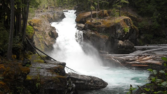 Aguas bravas fluyen sobre un cauce rocoso hacia un río azul.