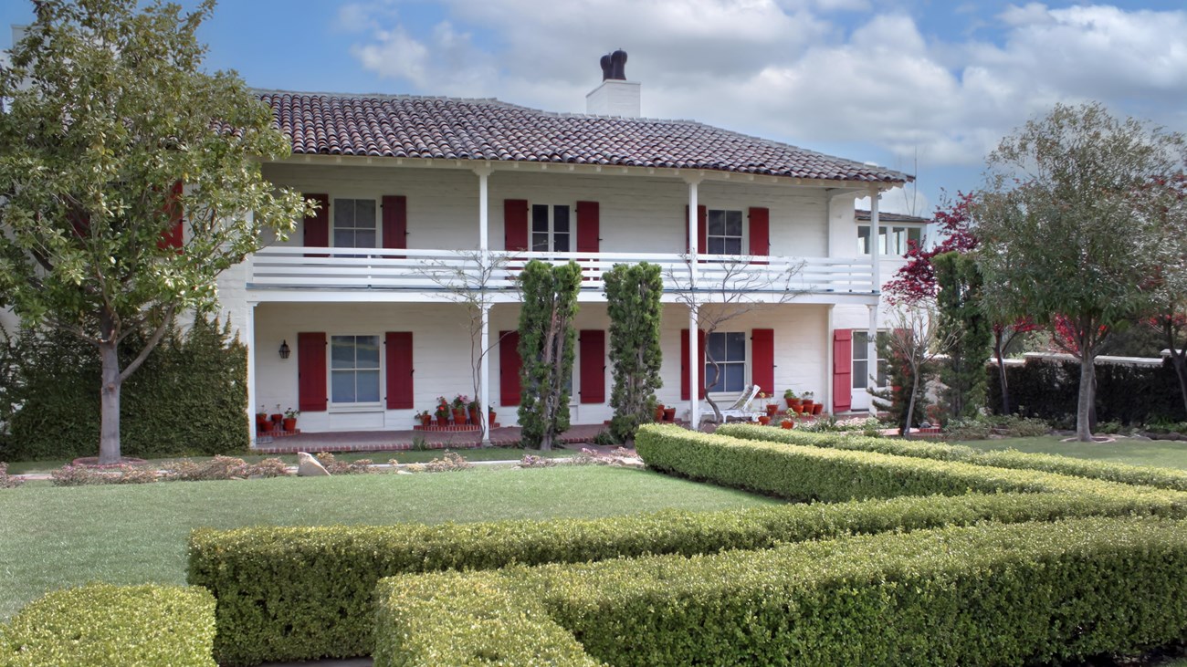 Hedges, trees and a courtyard are seen next to a two story home with shutters.