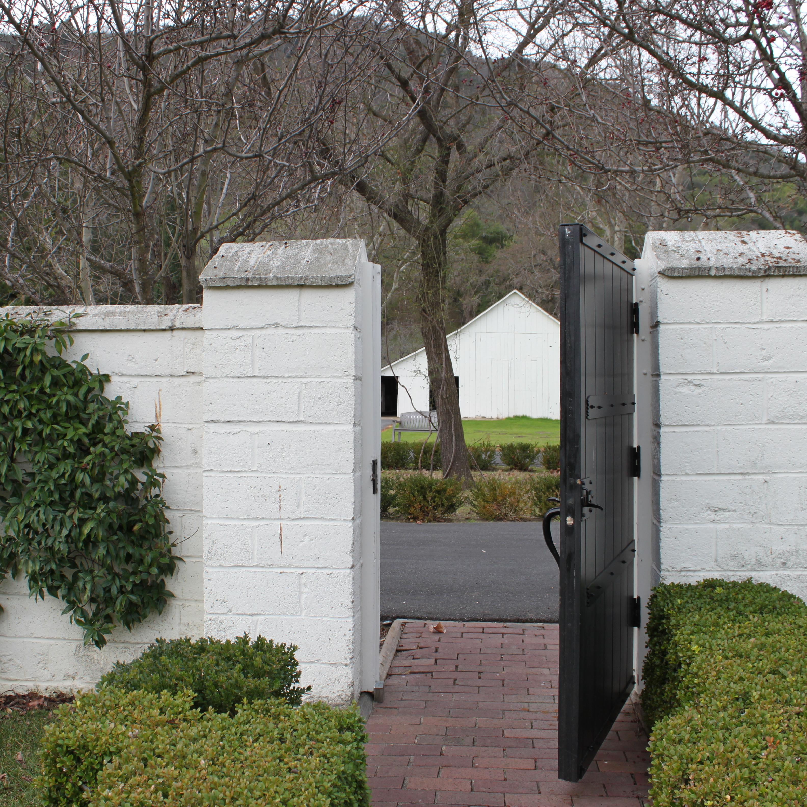 Photo of an entrance with white brick and a brown gate that's open. A barn is seen in the background