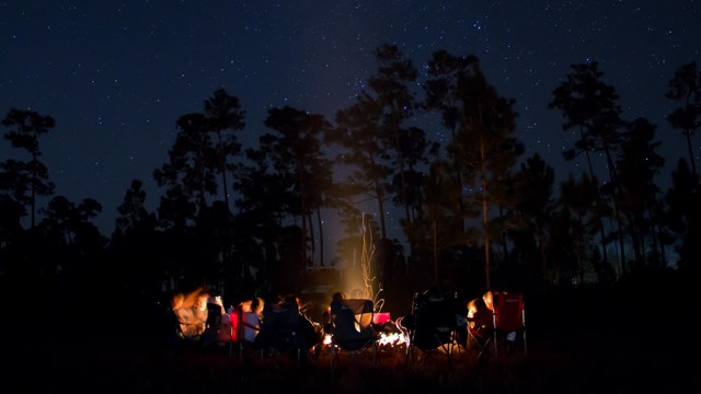 A group of people sit in chairs around a campfire under a night sky.