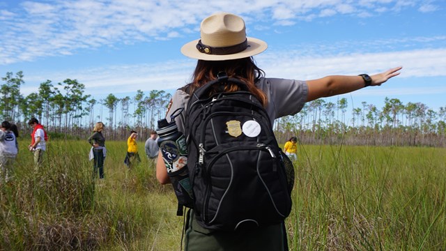 A ranger stands in vegetation and points right.