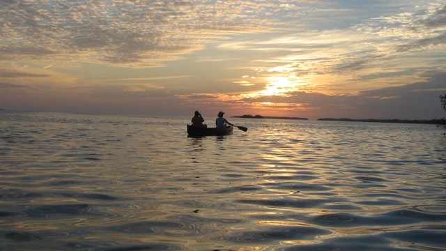 Two paddlers on a canoe floating on rippled water. The sun is setting in the background.