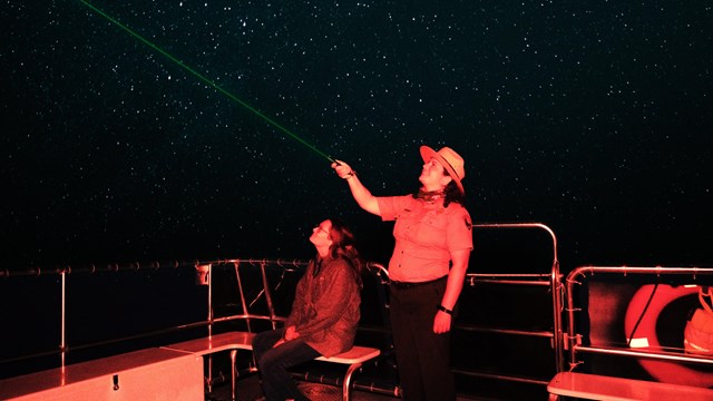 A Park Ranger stands on a boat while holding up a laser pointer to the night sky. A visitor sits nea