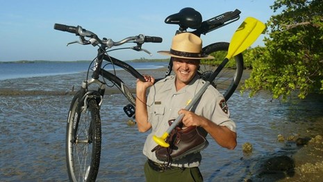 A park ranger standing by the water holds up a paddle and a bicycle. 