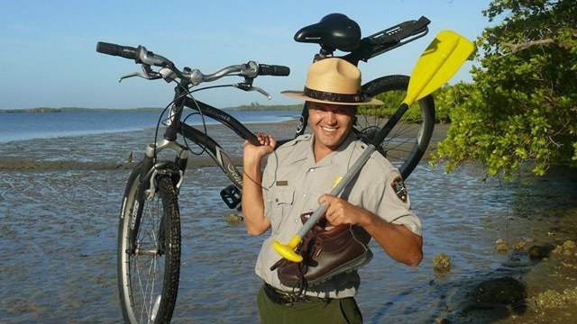 A park ranger standing by the water holds up a paddle and a bicycle. 