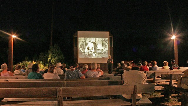 Visitors sit on benches at an evening slideshow program.
