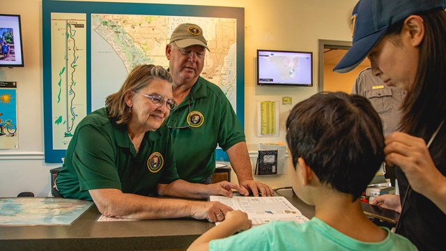 Two Park Volunteers behind a desk at the Visitor Center point to papers while helping a kid and thei