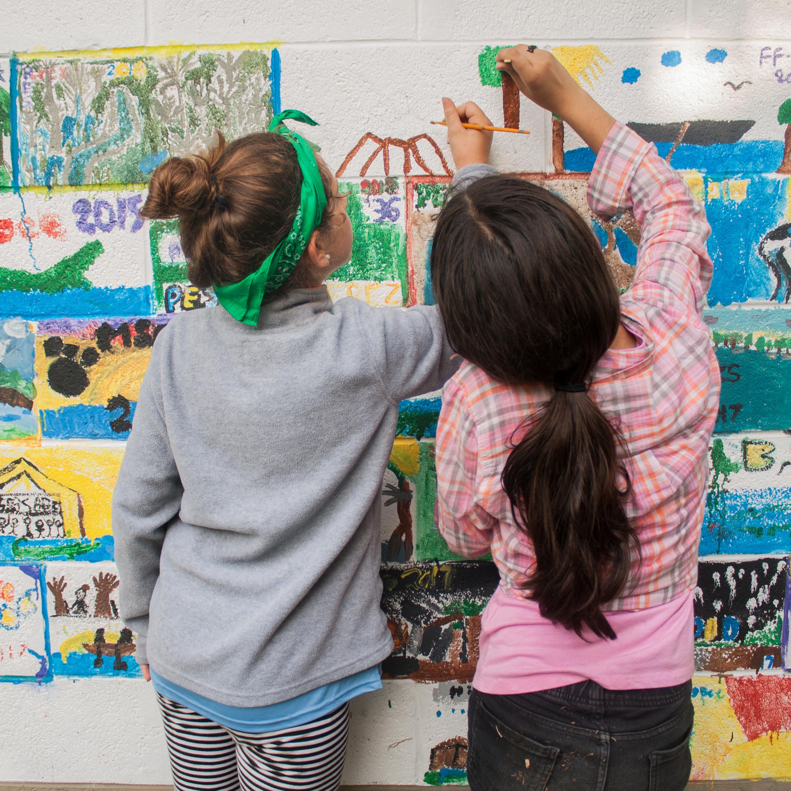Two girls painting a colorful mural on a wall with their backs towards the camera 