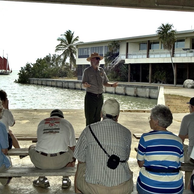 A visitor audience sits on benches listening to a male Park Ranger in green and gray uniform