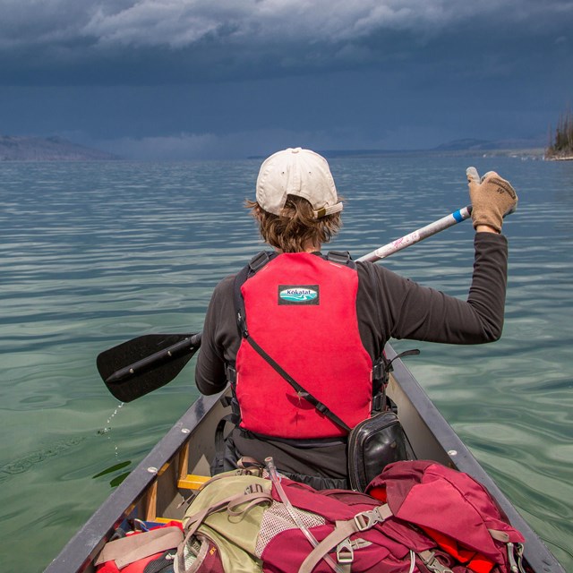 a boy in a red lifejacket paddles a canoe on a lake
