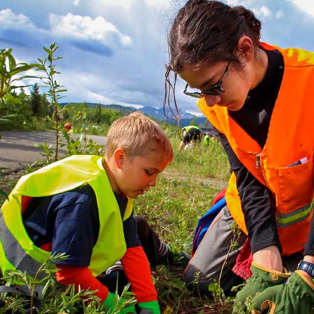 a young boy and girl in safety vests pull weeds