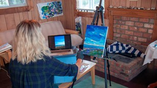 A woman paints on canvas in a room filled with artwork and supplies.