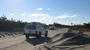 Two vehicles drive on sand road.