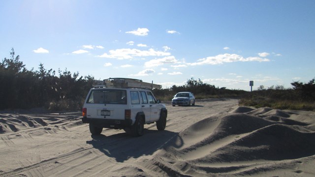 Two vehicles drive on sand road.