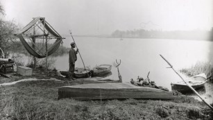 A black and white photograph features a gentleman on the shore of a river holding a fishing spear an