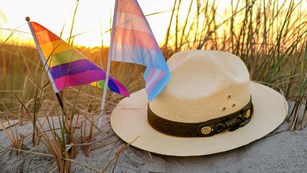 A park rangers flat hat sits atop a dune next to an LGBTQ pride flag and transgender pride flag.