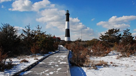 A snowy boardwalk leads to a black and white lighthouse tower.