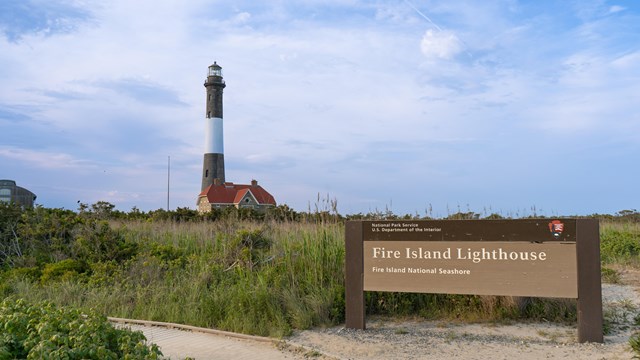 A sign reading "Fire Island Lighthouse" with a black and white striped lighthouse in the background.