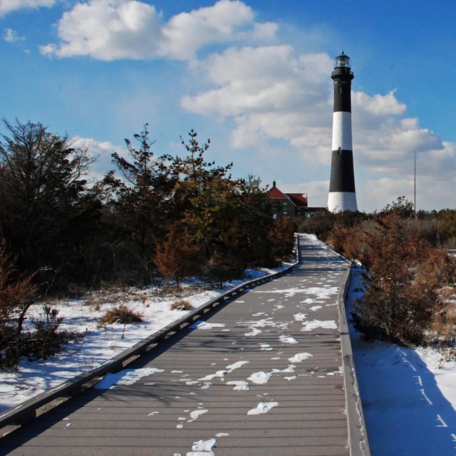 A snowy boardwalk leads to a black and white lighthouse tower.