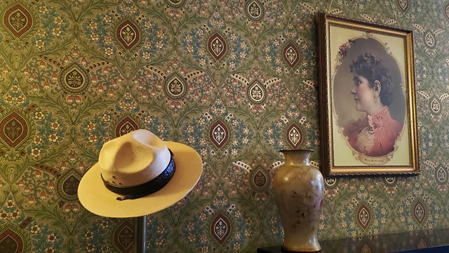 a flat hat sits on a stanchion in front of wall paper, next to a portrait of Ida McKinley