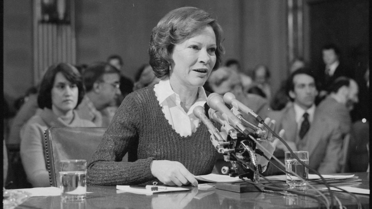 Rosalynn Carter pictured seated at a desk with several microphones pointed at her