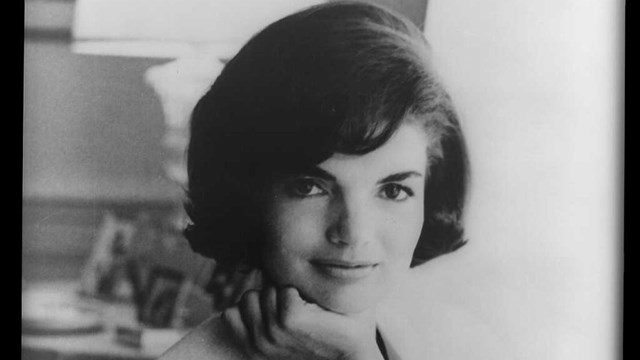 Jackie Kennedy sits with her arm draped over a couch and her other hand tucked up under her chin