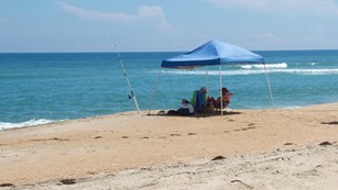 Visitors under a popup tent next to fishing rods planted in the sand