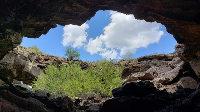 Looking up, a circular opening in a rock surface opens to a cloud-dotted sky above