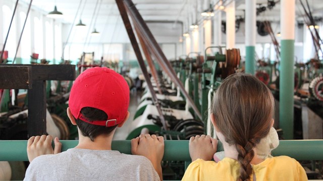 Two kids peering over a railing to look at weaving machines in a cotton mill