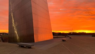Base of a tall metal structure on a plaza turning red with the sunset