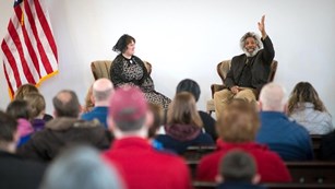 Two reenactors sit on a stage in front of an audience.